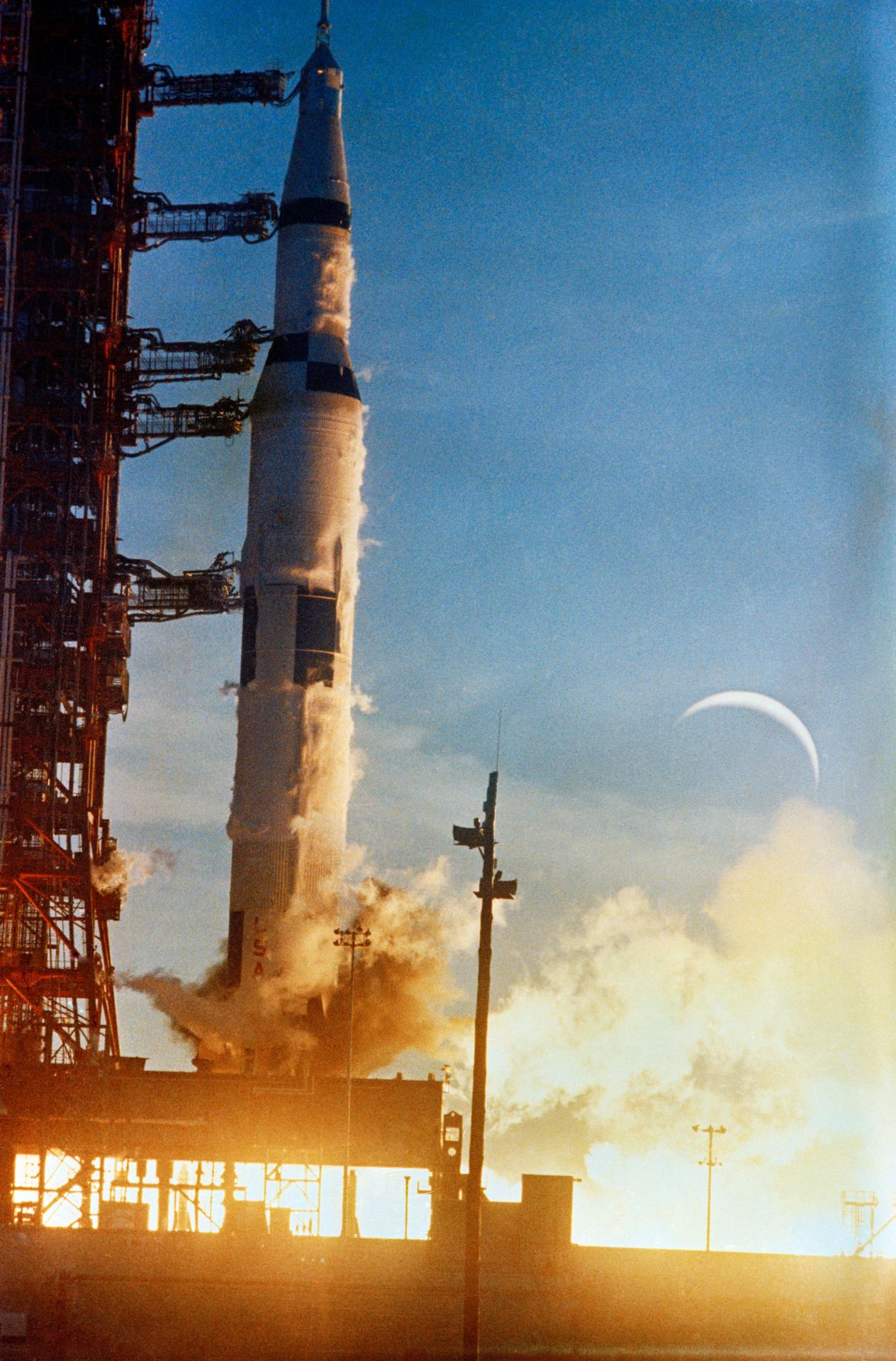 The Apollo 8 Saturn V an instant after ignition at dawn, engines lit and venting, still beside the launch tower, with a thin crescent moon in the sky.
